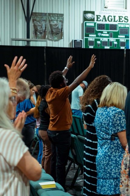 A man with both arms raised in worship, the gym scoreboard behind him