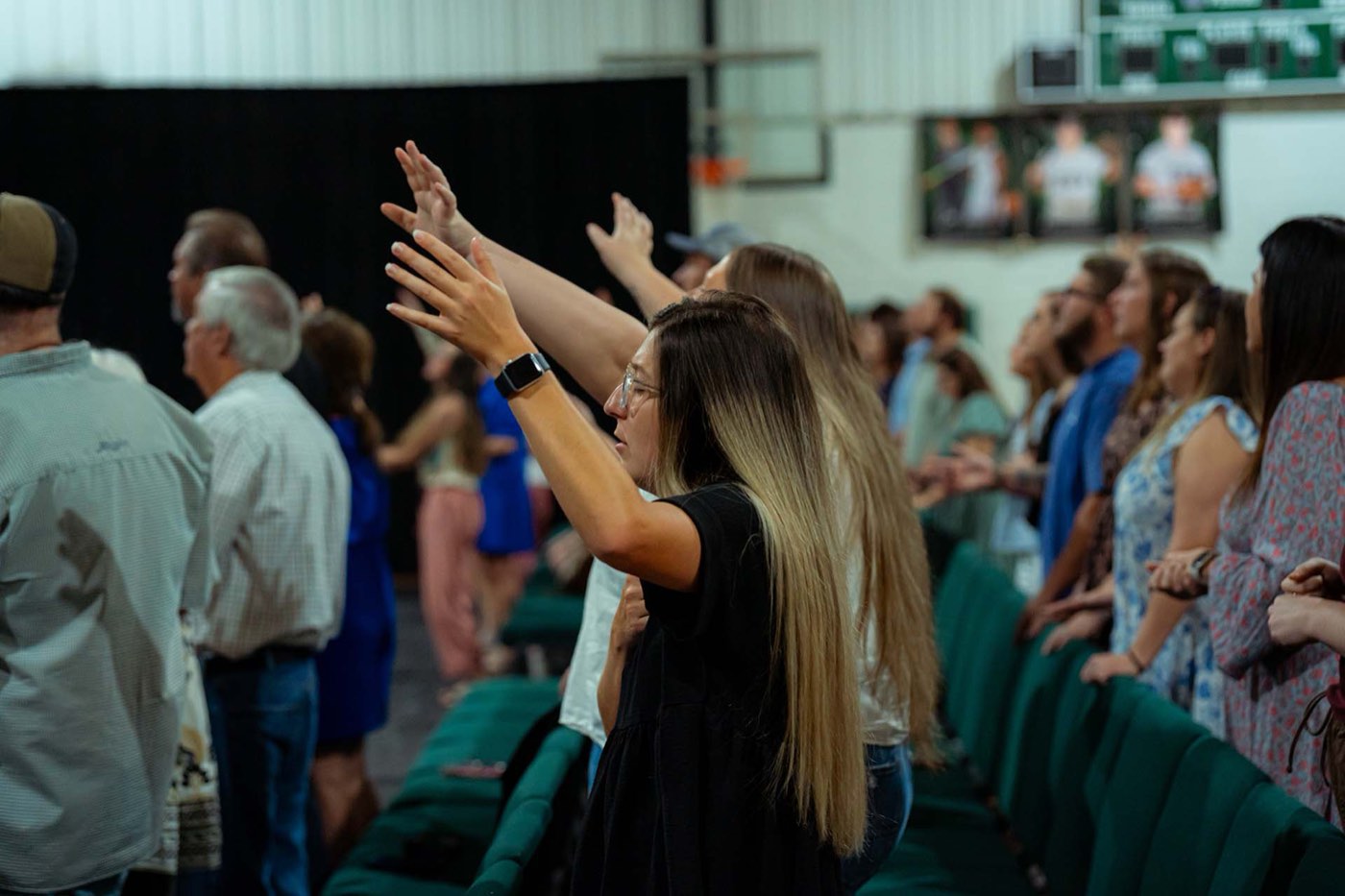 A woman with both arms raised during worship, rows of people singing behind her